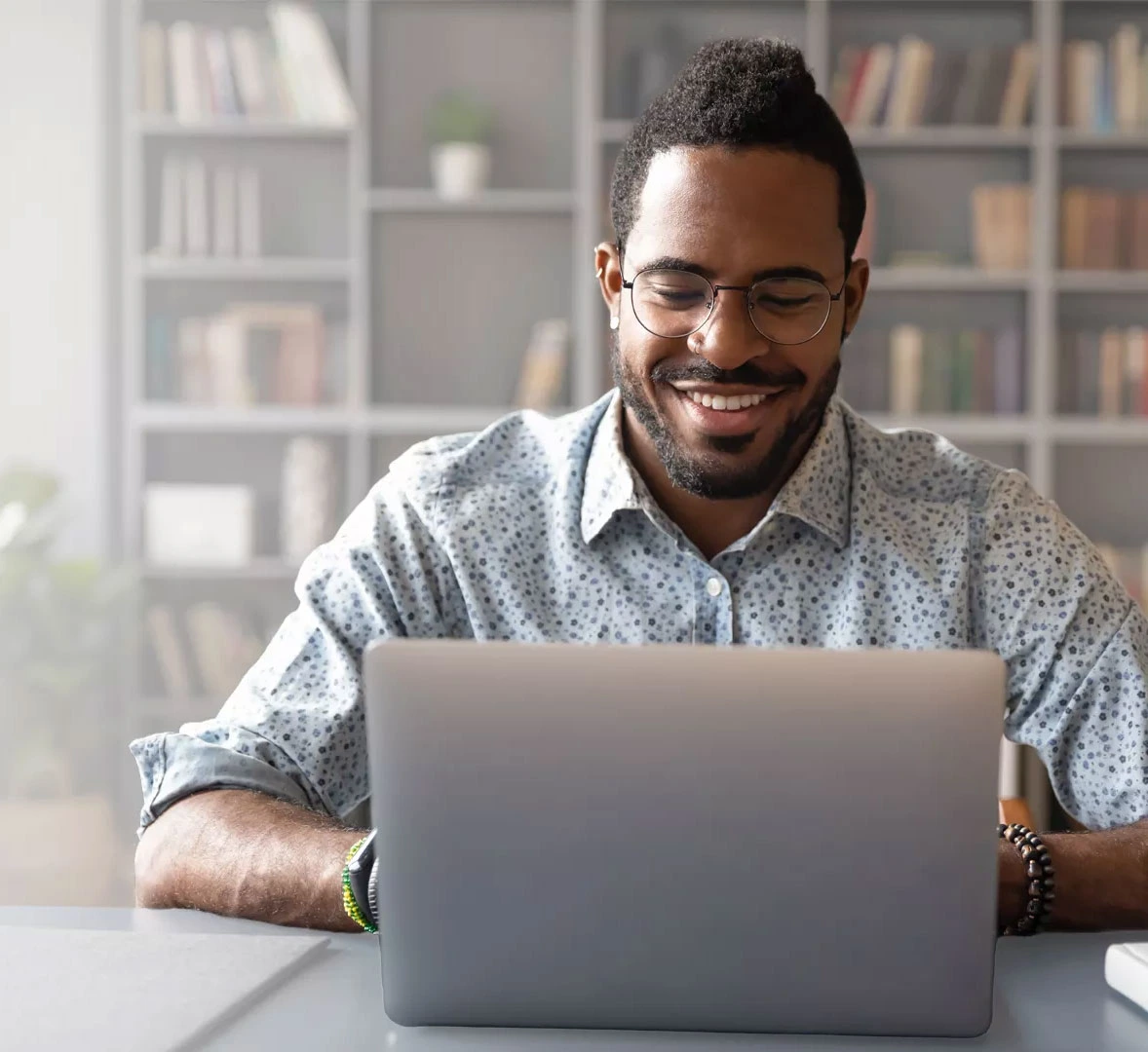 A smiling man wearing glasses is focused on his laptop, engaged in work with a positive demeanor.
